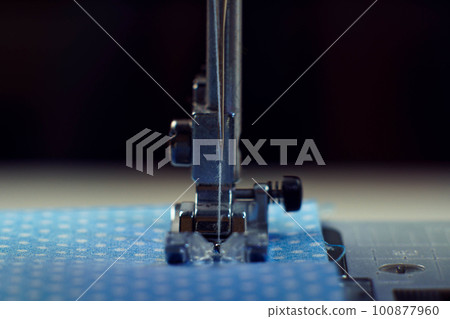 Detailed close up shot of sewing machine steel foot with needle and thread stitching on blue fabric polka dot textile 100877960