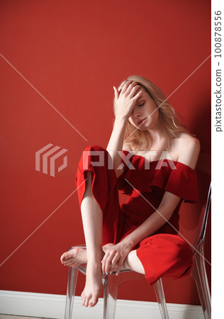 Beautiful young adult woman wearing red jumpsuit relaxing on chair 100878556