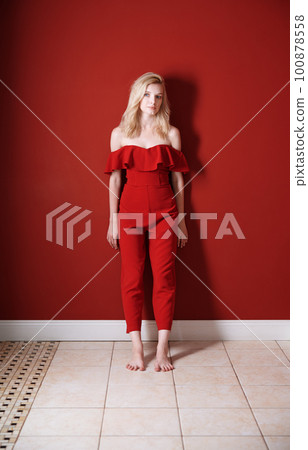 Beautiful young adult woman wearing red jumpsuit standing next to the wall Beautiful young adult woman wearing red jumpsuit standing next to the wall 100878558
