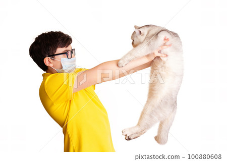 A boy in a medical mask holds his white cat at arms length 100880608