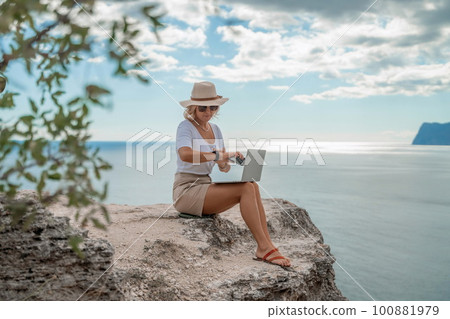 Freelance women sea working on the computer. Good looking middle aged woman typing on a laptop keyboard outdoors with a beautiful sea view. The concept of remote work. 100881979