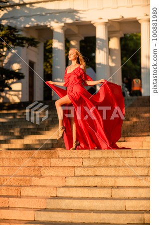 A woman in a long red dress against the backdrop of sunrise, bright golden light of the sun's rays. The concept of femininity, harmony. 100881980