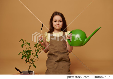 Adorable smiling little kid girl holding watering can and garden shovel, learns gardening, planting and cultivating vegetables in domestic garden, isolated on beige backdrop. Ecology, organic food. Adorable smiling little kid girl holding watering can and garden shovel, learns gardening, planting and cultivating vegetables in domestic garden, isolated on beige backdrop. Ecology, organic food. 100882506