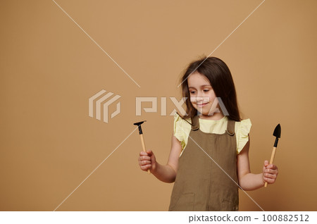 Adorable little toddler girl holds small garden tools - shovel and rake on beige background. Cute child learns gardening, planting and cultivating organic vegetables. Children and nature. Environment Adorable little toddler girl holds small garden tools - shovel and rake on beige background. Cute child learns gardening, planting and cultivating organic vegetables. Children and nature. Environment 100882512