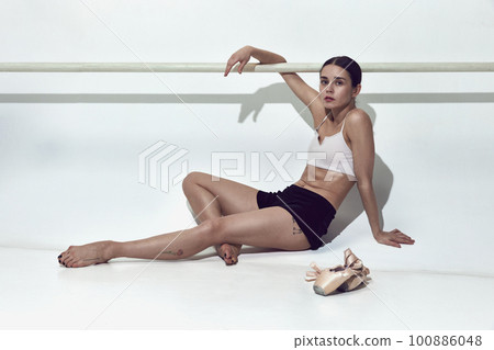 Tired beautiful ballerina sitting near dance bar and resting after hard work in dance hall over white studio background Tired beautiful ballerina sitting near dance bar and resting after hard work in dance hall over white studio background 100886048