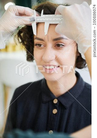 Esthetician using measuring tool for brow correction procedure in beauty salon. Close up. Beauty specialist placing ruler on smiling woman forehead while taking measurements for eyebrow correction. 100886074