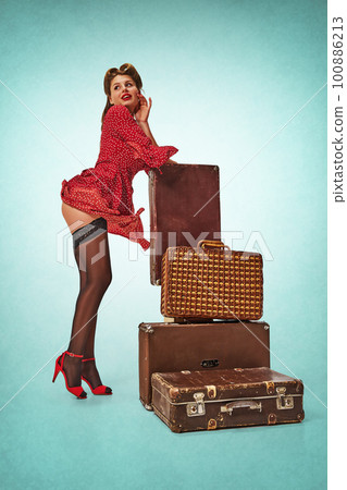 Portrait of sexy young girl in attractive red dress and stockings standing near vintage suitcases against blue background. Treveller Portrait of sexy young girl in attractive red dress and stockings standing near vintage suitcases against blue background. Treveller 100886213