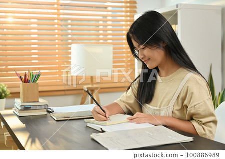 Cute asian teenage student, doing homework, writing task, taking notes on desk at home Cute asian teenage student, doing homework, writing task, taking notes on desk at home 100886989
