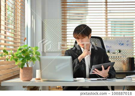 Smart young asian male economist holding digital tablet and analyzing financial data, stock market information on laptop computer 100887018