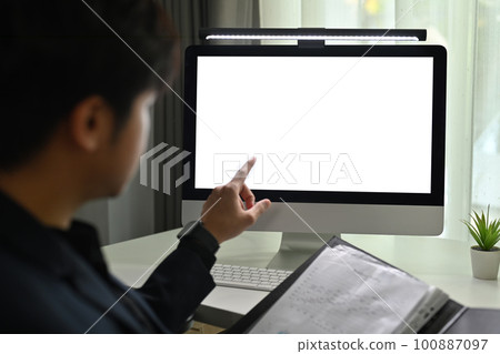 Back view of young businessman holding open office binder and pointing fingers at white blank screen computer monitor Back view of young businessman holding open office binder and pointing fingers at white blank screen computer monitor 100887097