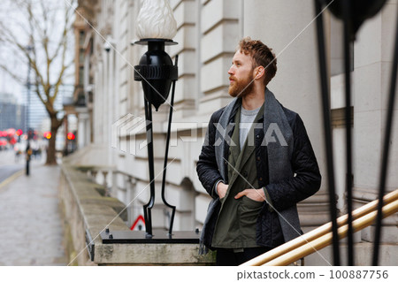 Stylishly dressed bearded man looks around standing near the entrance to a large building 100887756