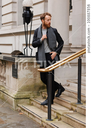 Stylish bearded man in a jacket and scarf descends the stairs leaving the building 100887856