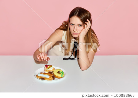 Upset young model surrounded by junk food looking at camera with sad eyes over pink background 100888663