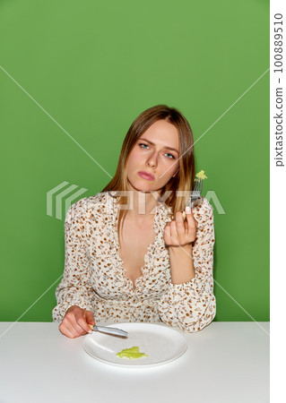 Portrait of exhausted young woman with peace of salad on plate and eating over green background 100889510