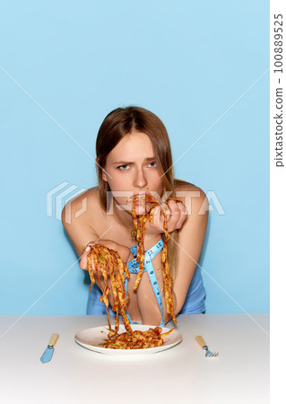 Portrait of adorable young girl eating spaghetti by hands with sad facial expression over blue background 100889525