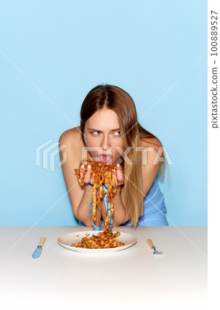 One young pretty girl with greedy face eating noodles by hands over blue background. Cheat meal, eating disorder concept 100889527