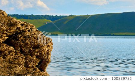 Part of the brown rock on the bank of a summer calm river with green hills Part of the brown rock on the bank of a summer calm river with green hills 100889785