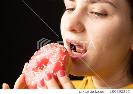 A woman bites a large red donut, a black background, a place for text. Gluttony, overeating and sugar addict. 100892225