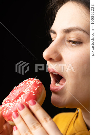 A woman bites a large red donut, a black background, a place for text. Gluttony, overeating and sugar addict. 100892228