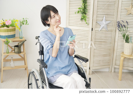 A woman in a wheelchair imagining while using a smartphone in the living room 100892777