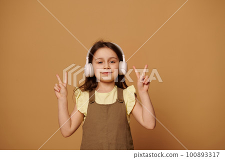 Caucasian cheery little child girl 5-6 years old, wearing wireless headphones, showing peace sign with her fingers, expressing positive emotions, smiling looking at camera, isolated beige background 100893317