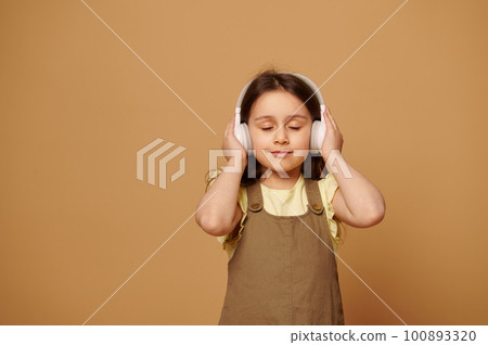 Happy relaxed little toddler girl listens to soundtrack on white wireless headphones, enjoys soothing music, posing with her eyes closed over isolated beige background with copy advertising space 100893320