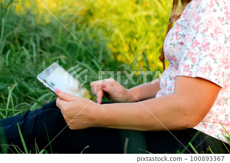Holding tablet outside. Young woman using tablet outdoor outside. Green grass in park on sunny summer day sitting on grass. Out of focus 100893367