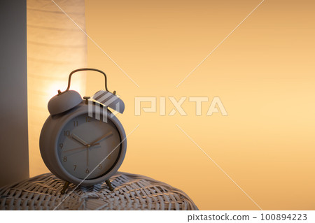 On the wicker lid of the basket stands an analog alarm clock against a background of a lit bedside lamp. 100894223