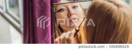 Young woman does makeup sitting by the window with a panoramic view of the skyscrapers and the big city BANNER, LONG FORMAT 100896028