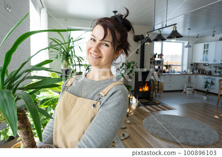 A happy woman in a green house with a potted plant in her hands smiles, takes care of a flower. The interior of a cozy eco-friendly house, a fireplace stove, a hobby for growing and breeding homeplant 100896103
