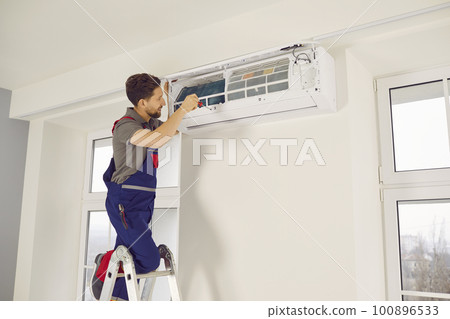 Professional electrician man with screwdriver maintaining modern air conditioner indoors. Professional electrician man with screwdriver maintaining modern air conditioner indoors. 100896533