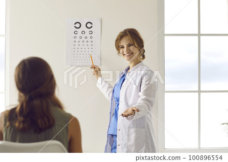 Happy Caucasian female optician in white medical uniform tests vision using Optometry table in optics salon. Happy Caucasian female optician in white medical uniform tests vision using Optometry table in optics salon. 100896554