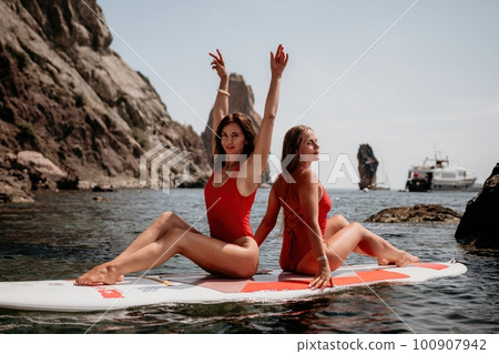 Woman sup yoga. Two happy sporty women practising yoga pilates on paddle sup surfboard. Female stretching doing workout on sea water. Modern individual female hipster outdoor summer sport activity. 100907942