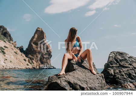 Woman summer travel sea. Happy tourist enjoy taking picture outdoors for memories. Woman traveler posing on the beach at sea surrounded by volcanic mountains, sharing travel adventure journey 100907944