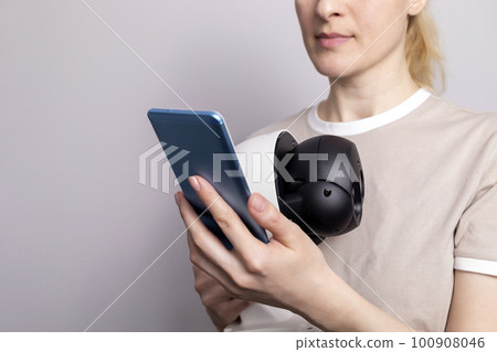 White Female Person Holds IP Wifi Outdoor Security Camera And Looks at Smartphone On White Grey Background. Smart Home, House Surveillance, Protection. Horizontal, closeup 100908046