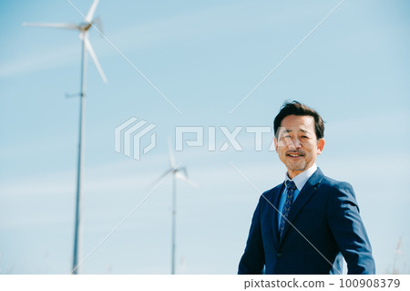 A businessman standing with a wind power generation facility in the background A businessman standing with a wind power generation facility in the background 100908379