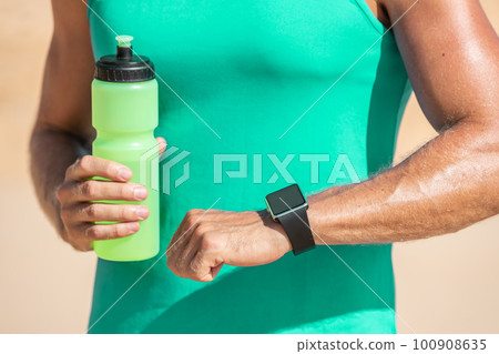 Sports smart watch athlete man running checking heart rate during cardio run workout break drinking water bottle on outdoor beach 100908635