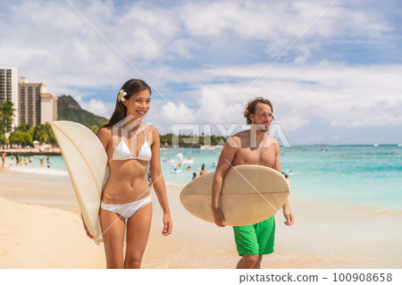 Hawaii Waikiki beach couple tourists surfers happy going surfing. Interracial couple, Asian bikini model woman, Caucasian man walking with surfboards after surf lesson 100908658
