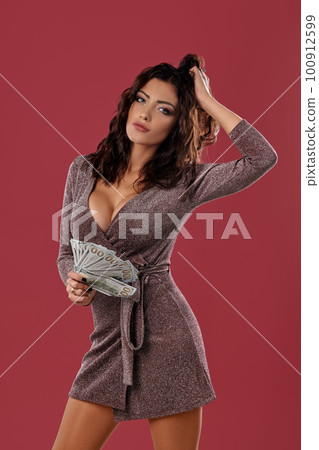 Brunette girl in shiny dress posing holding a fan of hundred dollar bills in her hand, standing on pink studio background. Casino, poker. Close-up. Brunette girl in shiny dress posing holding a fan of hundred dollar bills in her hand, standing on pink studio background. Casino, poker. Close-up. 100912599