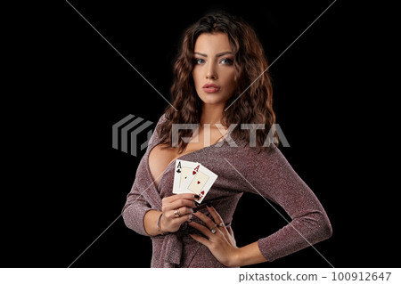 Brunette girl wearing shiny dress posing holding two playing cards in her hand standing against black studio background. Casino, poker. Close-up. 100912647