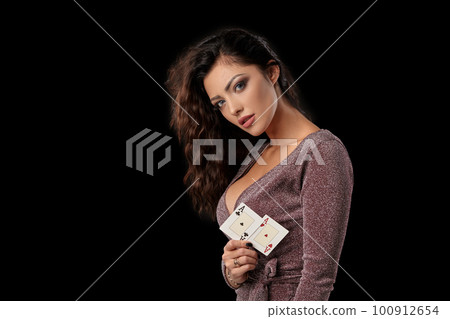 Brunette girl wearing shiny dress posing holding two playing cards in her hand standing against black studio background. Casino, poker. Close-up. Brunette girl wearing shiny dress posing holding two playing cards in her hand standing against black studio background. Casino, poker. Close-up. 100912654