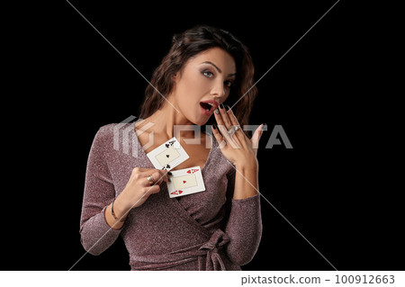 Brunette girl wearing shiny dress posing holding two playing cards in her hand standing against black studio background. Casino, poker. Close-up. 100912663