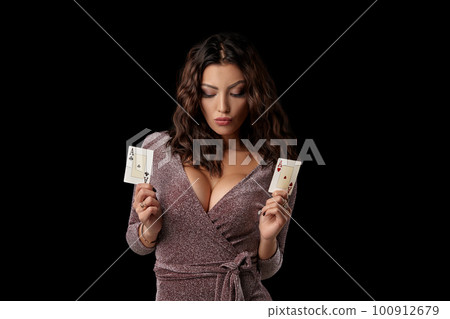 Brunette girl wearing shiny dress posing holding two playing cards in her hands standing against black studio background. Casino, poker. Close-up. 100912679