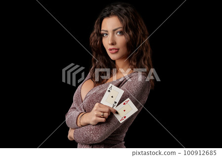 Brunette girl wearing shiny dress posing holding two playing cards in her hands standing against black studio background. Casino, poker. Close-up. Brunette girl wearing shiny dress posing holding two playing cards in her hands standing against black studio background. Casino, poker. Close-up. 100912685