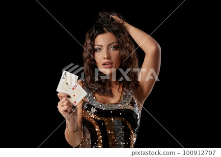 Brunette girl in a colored sequin dress posing holding two playing cards, standing against black studio background. Casino, poker. Close-up. Brunette girl in a colored sequin dress posing holding two playing cards, standing against black studio background. Casino, poker. Close-up. 100912707