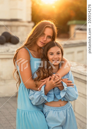 Portrait of mother and daughter in blue dresses with flowing long hair against the backdrop of sunset. The woman hugs and presses the girl to her. They are looking at the camera. 100914276