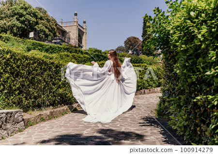 Brunette runs white dress park. A beautiful woman with long brown hair and a long white dress runs along the path along the beautiful bushes in the park, rear view Brunette runs white dress park. A beautiful woman with long brown hair and a long white dress runs along the path along the beautiful bushes in the park, rear view 100914279