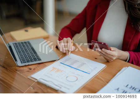 Portrait of a young Asian woman showing a smiling face as she uses her phone, computer and financial documents on her desk in the early morning hours. Portrait of a young Asian woman showing a smiling face as she uses her phone, computer and financial documents on her desk in the early morning hours. 100914693