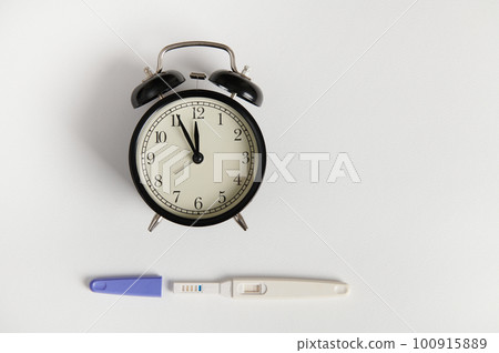 Flat lay with black retro alarm clock and pregnancy test kit showing positive result, isolated white backdrop. Pregnancy, motherhood and gynecological concept. Women's health and fertility. Still life 100915889