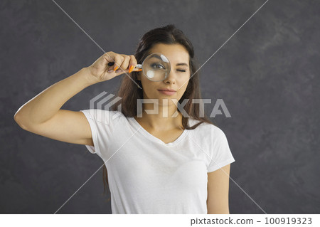 Close up studio portrait of young woman looking at camera through magnifying glass. Close up studio portrait of young woman looking at camera through magnifying glass. 100919323